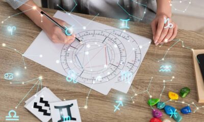 Mãos escrevendo em mapa astral com símbolos do zodíaco e pedras em mesa de madeira.