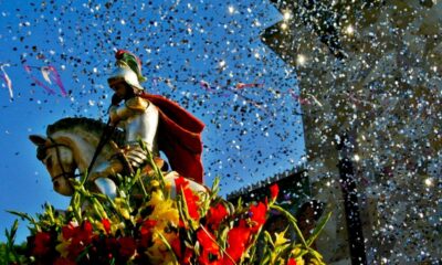 Estátua de São Jorge cavaleiro com armadura e capa vermelha, adornada com flores, sob chuva de confetes.
