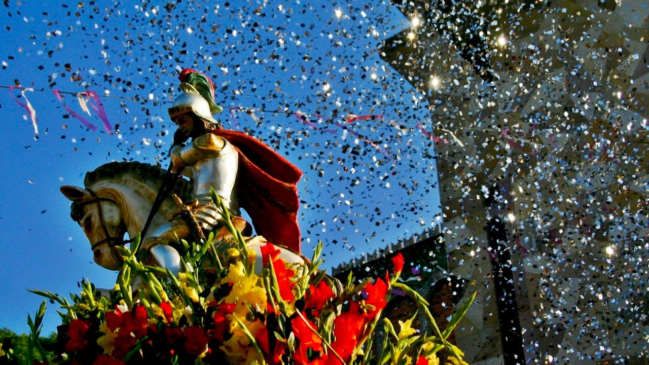 Estátua de São Jorge cavaleiro com armadura e capa vermelha, adornada com flores, sob chuva de confetes.