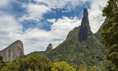 Picos de montanhas rochosas cobertos por floresta densa sob céu azul com nuvens brancas.
