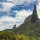 Picos de montanhas rochosas cobertos por floresta densa sob céu azul com nuvens brancas.