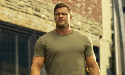 Homem musculoso com camiseta verde militar e barba por fazer, olhando sério, em fundo urbano.