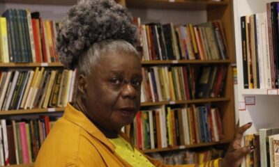 Mulher negra com cabelo grisalho crespo em livraria, tocando livro, olhando para câmera.