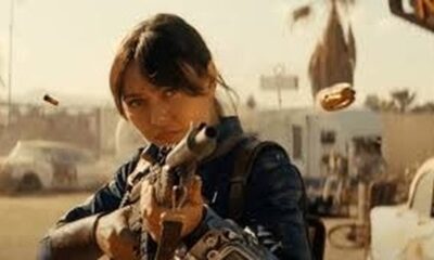 Mulher jovem com rifle mira em ambiente árido pós-apocalíptico com cápsula de bala no ar.