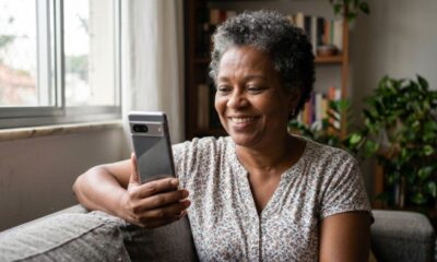 Mulher sorridente segura smartphone, olhando para a tela enquanto sentada em um sofá.