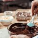 Pessoa mexendo chocolate derretido em tigela de vidro com espátula em ambiente de cozinha.