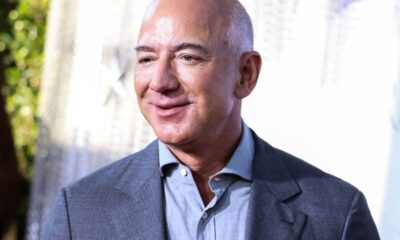 Jeff Bezos em close, sorrindo, usando camisa azul clara e blazer cinza escuro.