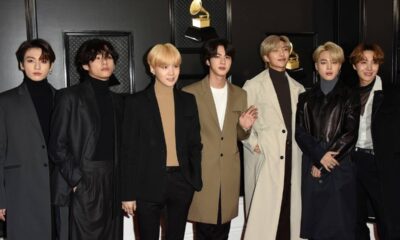 Sete membros do BTS, com casacos e golas rolê, posam em tapete vermelho com troféus.