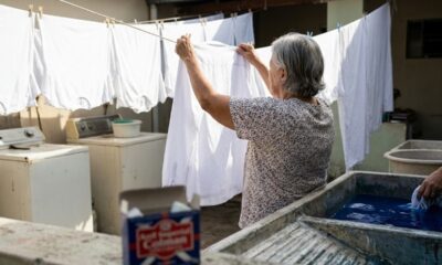 Mulher pendura camisa branca em varal; ao fundo, máquina de lavar e tanques com anil.