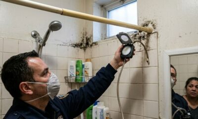 Homem mascarado inspeciona mofo preto em parede de chuveiro com aparelho; mulher observa no espelho.