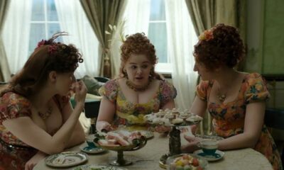 Três mulheres de Bridgerton, em trajes florais, desfrutam chá da tarde com doces e xícaras.