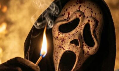 Máscara branca de Ghostface com sangue, capuz preto, segurando fósforo aceso em fundo flamejante.