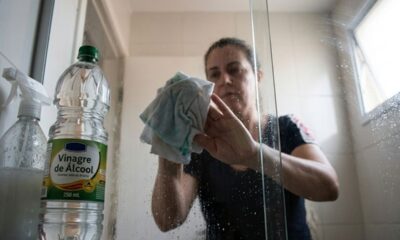 Mulher limpa box de banheiro com pano; garrafa de "Vinagre de Álcool" à esquerda.
