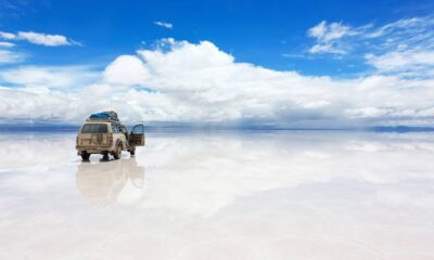 Jipe bege em salar refletindo céu azul com nuvens.