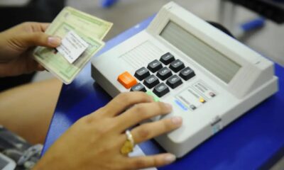Mãos de pessoa digitando em urna eletrônica e segurando documentos eleitorais.