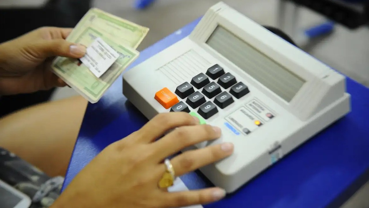 Mãos de pessoa digitando em urna eletrônica e segurando documentos eleitorais.