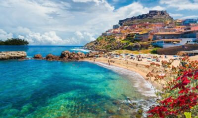 Vista aérea de praia com águas claras, rochas, vila colorida na colina e muitas pessoas.