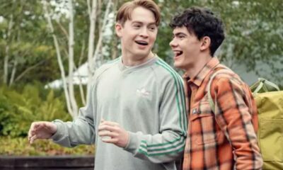 Dois jovens sorrindo, um de moletom cinza, outro de camisa xadrez laranja com mochila.