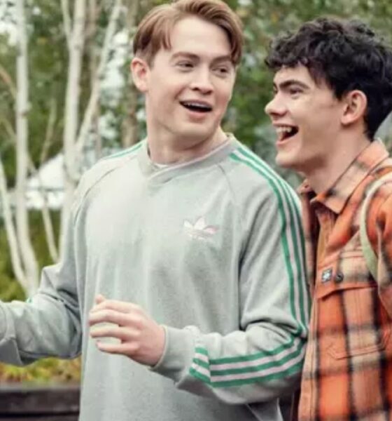 Dois jovens sorrindo, um de moletom cinza, outro de camisa xadrez laranja com mochila.