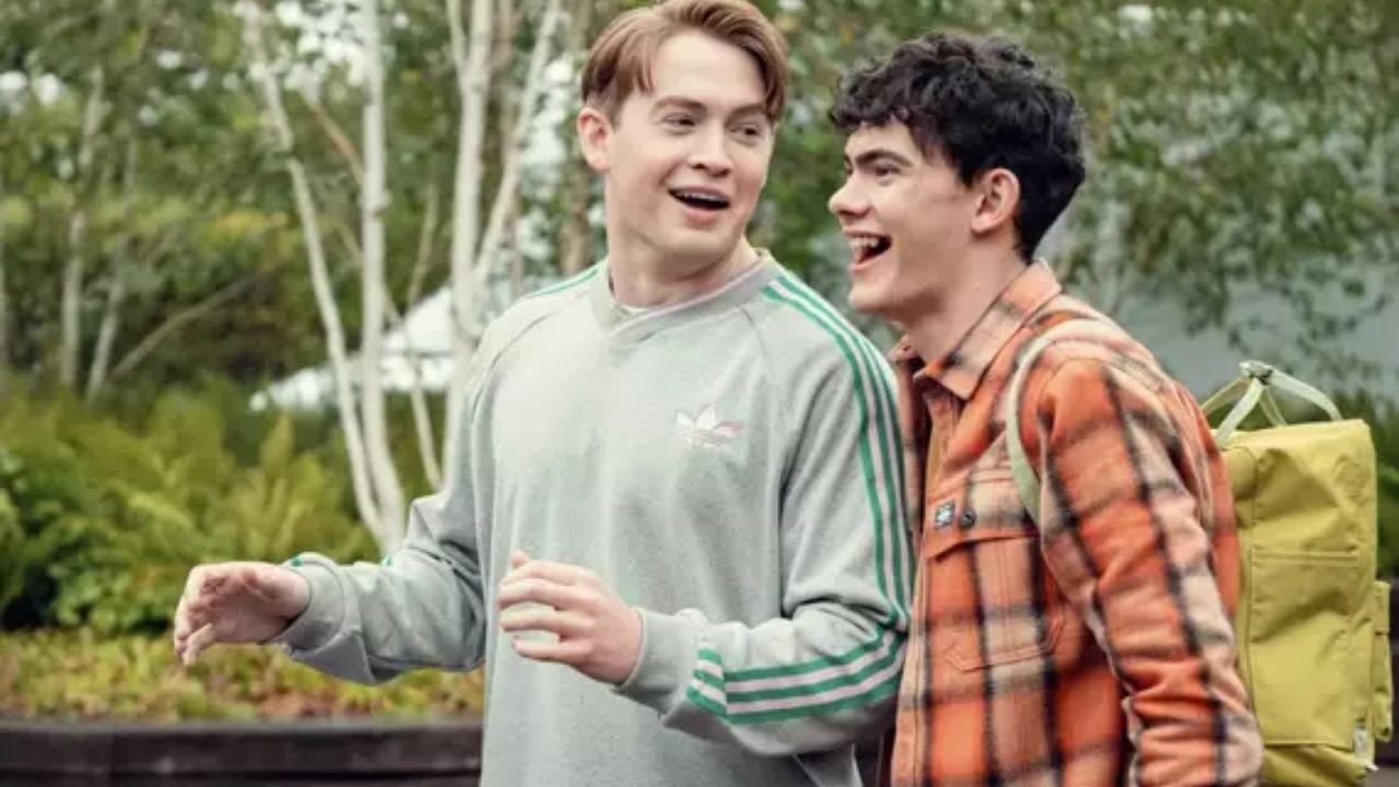 Dois jovens sorrindo, um de moletom cinza, outro de camisa xadrez laranja com mochila.