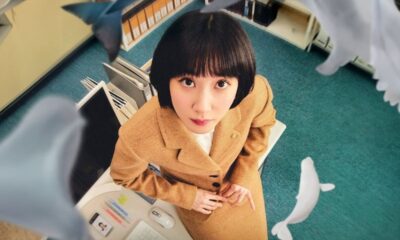 Mulher de blazer bege olhando para cima em escritório, com baleias brancas flutuando ao redor.