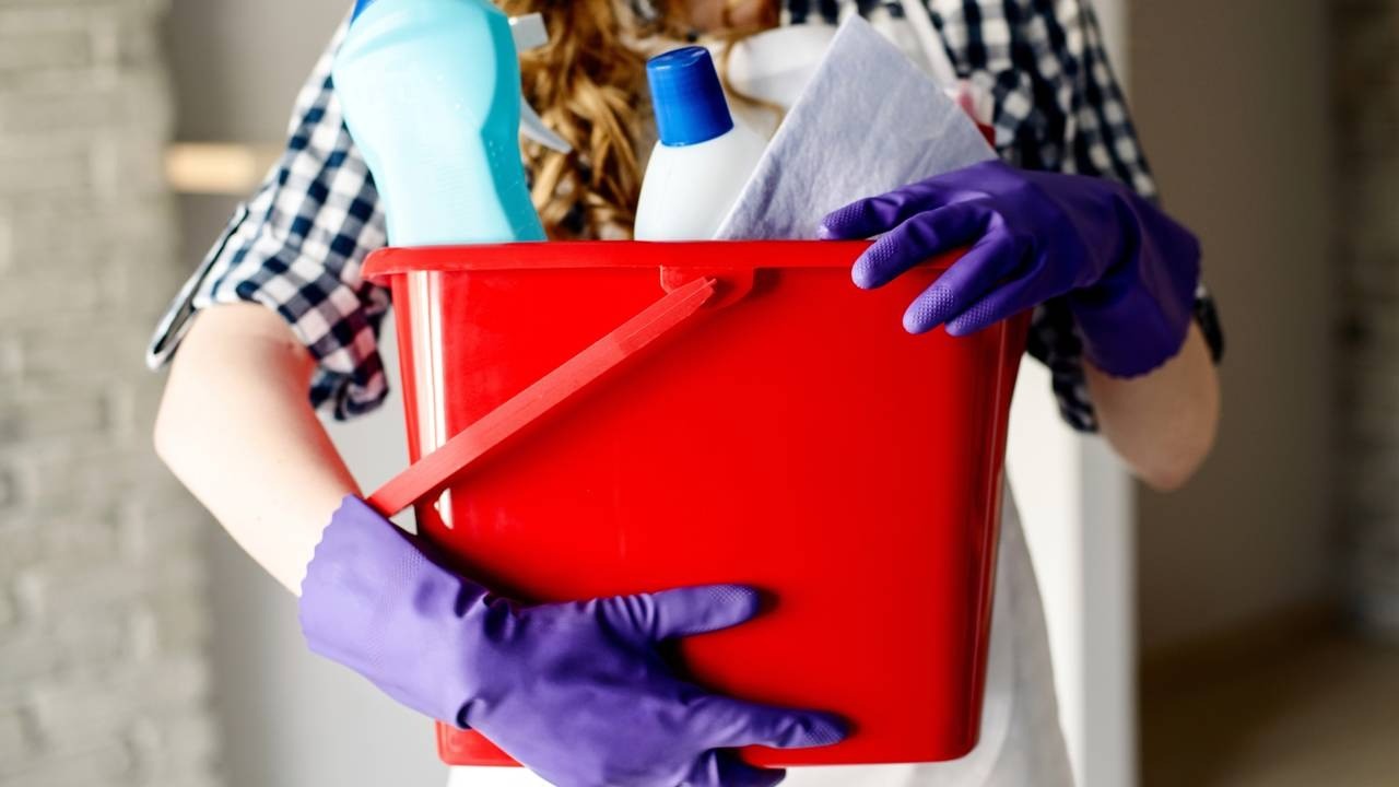 Pessoa com luvas roxas segurando balde vermelho com garrafas de produtos de limpeza e pano.