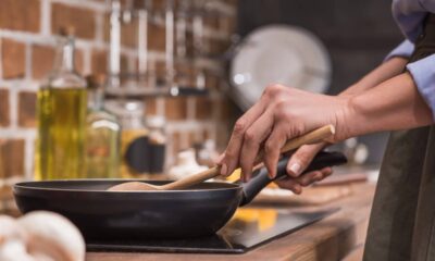Mãos seguram colher de pau em frigideira preta no fogão, cozinhando em cozinha com fundo de tijolos.