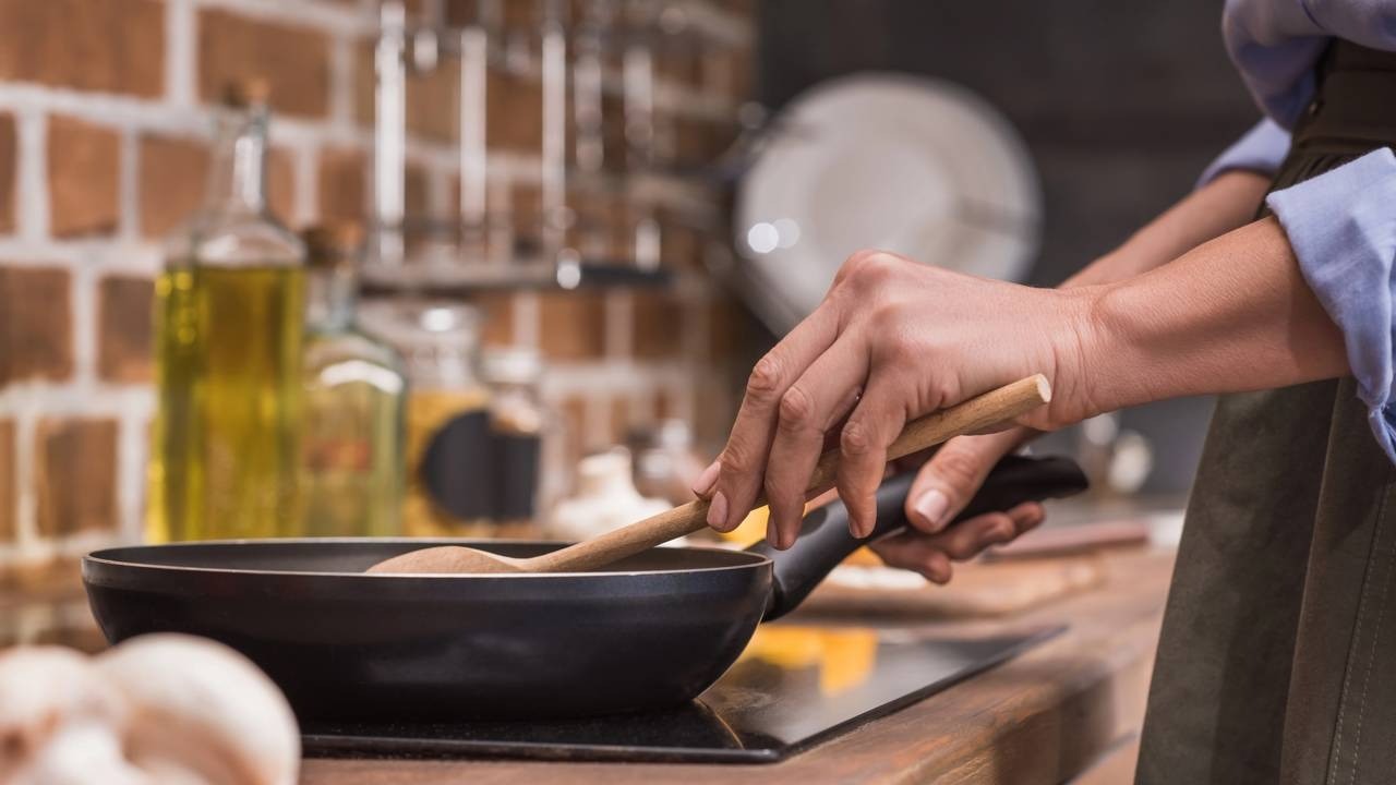 Mãos seguram colher de pau em frigideira preta no fogão, cozinhando em cozinha com fundo de tijolos.
