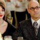 Stanley Tucci de óculos e Anne Hathaway de batom vermelho sentados em uma mesa.