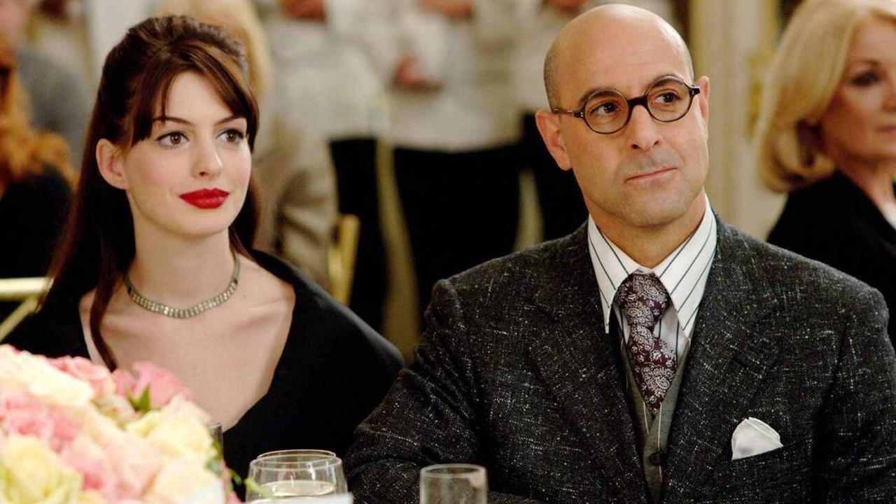 Stanley Tucci de óculos e Anne Hathaway de batom vermelho sentados em uma mesa.