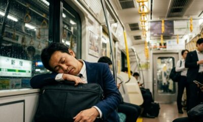 Homem de terno dorme em trem japonês chuvoso, apoiado em pasta, com letreiro 'Shinjuku'.