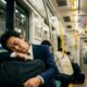 Homem de terno dorme em trem japonês chuvoso, apoiado em pasta, com letreiro 'Shinjuku'.