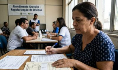 Mulher escrevendo formulário em atendimento de regularização de dívidas, com banner ao fundo.