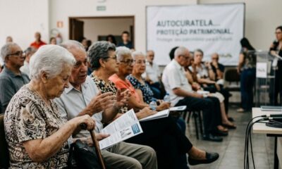 Idosos assistem a uma palestra sobre autocuratela e proteção patrimonial, com banner visível ao fundo.