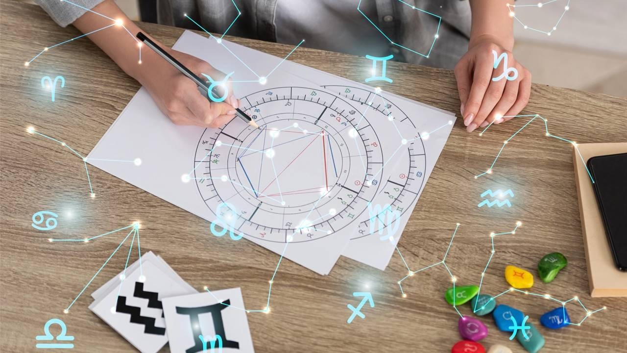 Mãos escrevendo em mapa astral com símbolos do zodíaco, cartas e pedras sobre mesa de madeira.