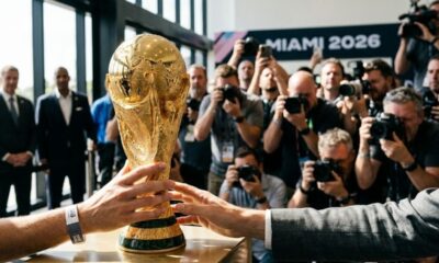 Mãos seguram o troféu da Copa do Mundo FIFA. Fotógrafos e banner 'MIAMI 2026' no fundo.
