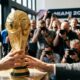 Mãos seguram o troféu da Copa do Mundo FIFA. Fotógrafos e banner 'MIAMI 2026' no fundo.