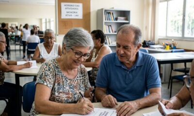 Mulher idosa sorri escrevendo, homem observa, outros idosos. Placa: 'MEMÓRIA ATIVA'.