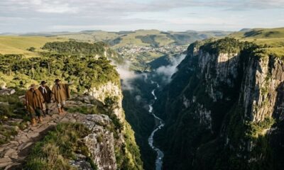 Três homens em trajes gaúchos em trilha rochosa, observando cânion vasto com rio e cidade.