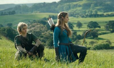 Jovem loiro e jovem mulher morena com arco, agachados em um campo verde.