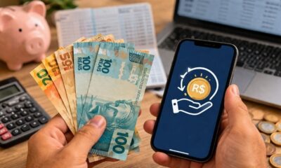 Mãos seguram notas de reais e um celular com ícone de resgate financeiro. Calculadora e cofrinho.