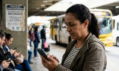 Mulher em ponto de ônibus urbano usando smartphone, outras pessoas também olham celulares.