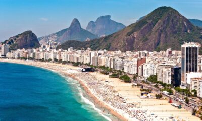 Vista aérea panorâmica da praia de Copacabana lotada, edifícios, mar azul e montanhas verdes.