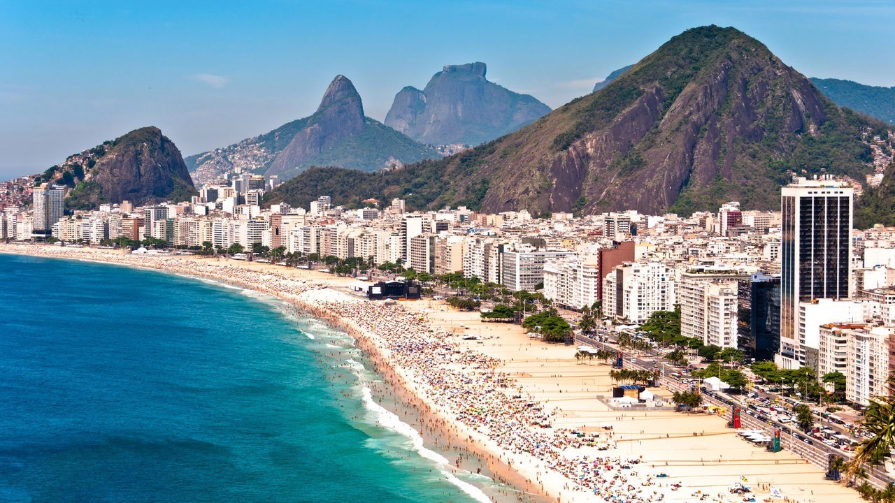 Vista aérea panorâmica da praia de Copacabana lotada, edifícios, mar azul e montanhas verdes.