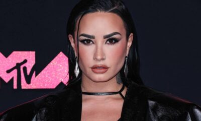Demi Lovato em close-up, maquiagem destacada, piercing no nariz, brincos, jaqueta preta. Fundo MTV.