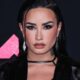 Demi Lovato em close-up, maquiagem destacada, piercing no nariz, brincos, jaqueta preta. Fundo MTV.