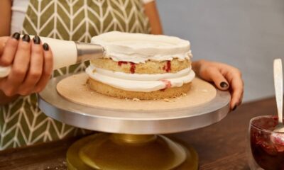 Mãos decoram bolo de camadas com glacê branco e recheio vermelho em suporte de confeitaria.