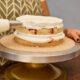Mãos decoram bolo de camadas com glacê branco e recheio vermelho em suporte de confeitaria.