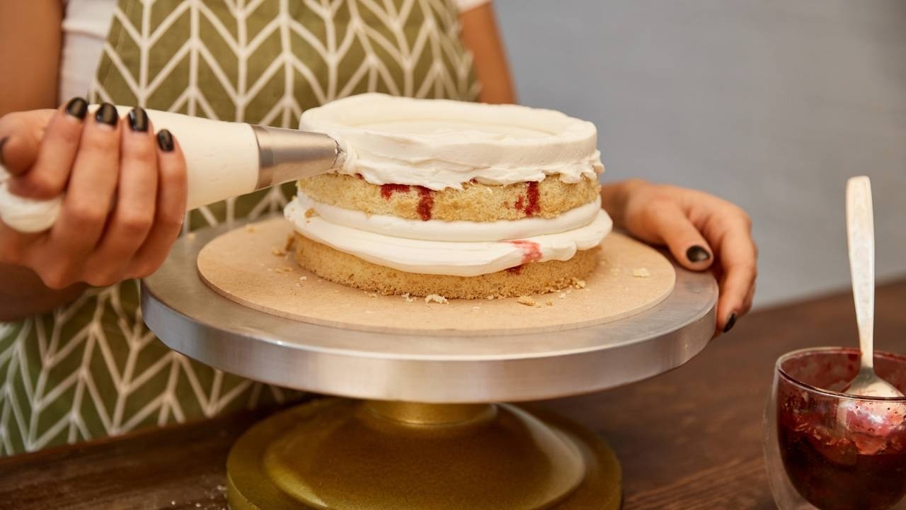 Mãos decoram bolo de camadas com glacê branco e recheio vermelho em suporte de confeitaria.