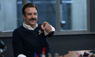 Jason Sudeikis como Ted Lasso sorrindo e apontando, vestindo suéter azul com brasão.